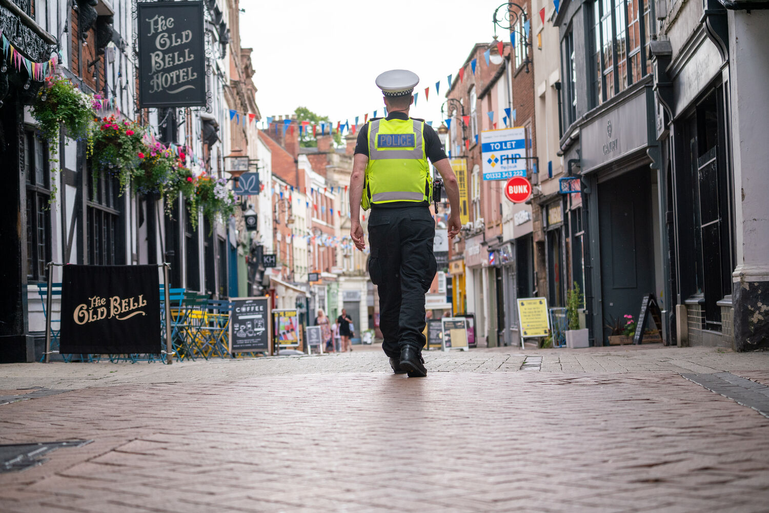 Officer in high visibility vest and uniform walking down city street with bar on the left and shops on the right