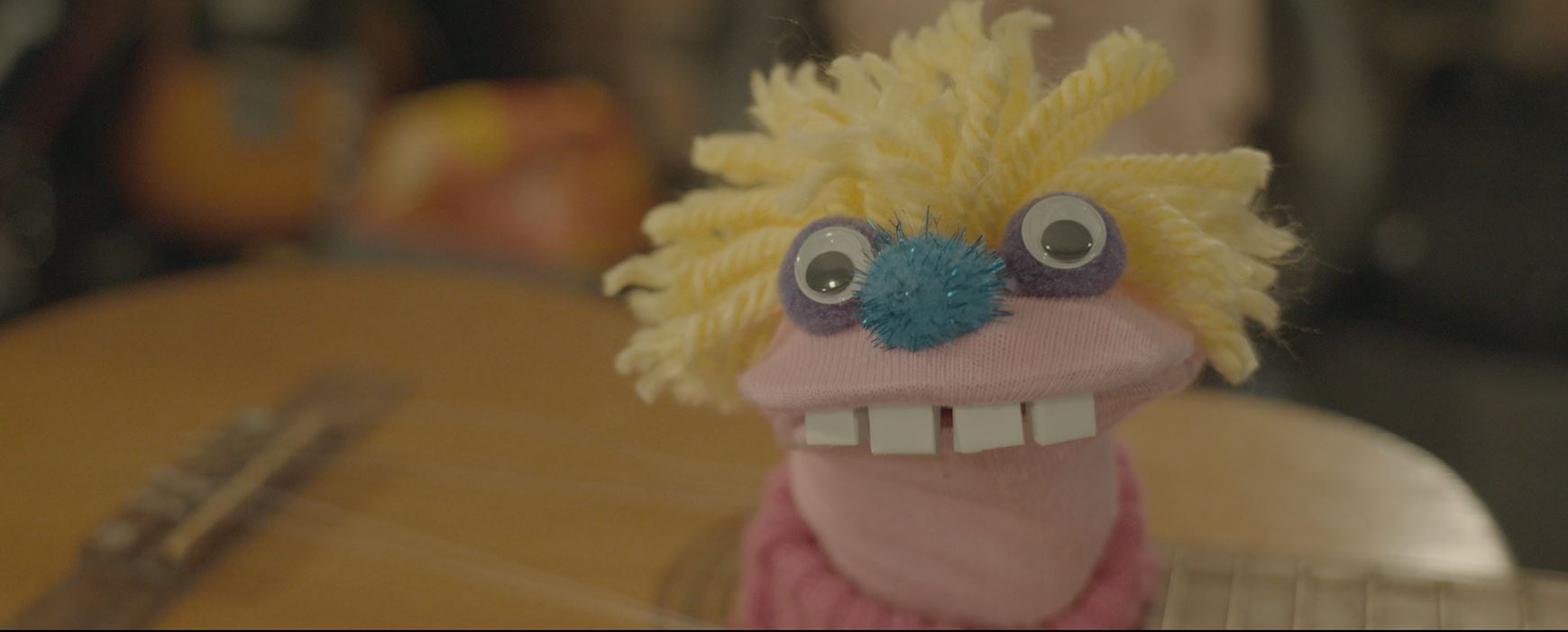 A bright pink sock puppet, with yellow hair and a blue pom pom nose looks at the camera stood in a guitar.