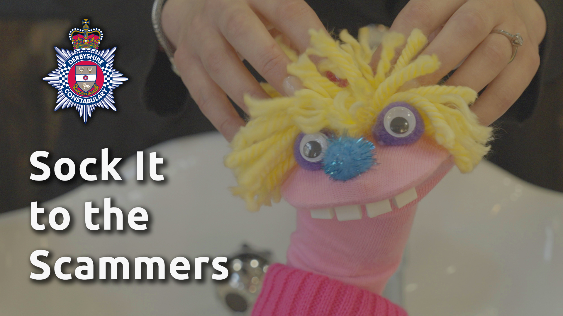 Claude the pink fraud puppet it having his bright yellow hair shampooed in a hair salon. He's loving it. Next to him is the Derbyshire Constabulary crest and the words "Sock It to the Scammers"