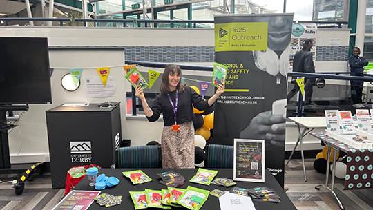 Beth Warrilow holding two flyers and standing in front of the 1625 banner at their stall in Derby Uni atrium