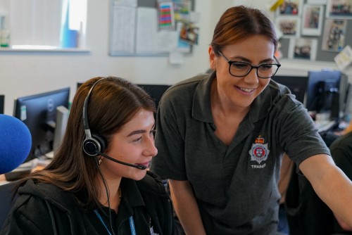 Female call handler, in uniform with dark brown hair, being assisted by another female colleague in uniform with blonde hair
