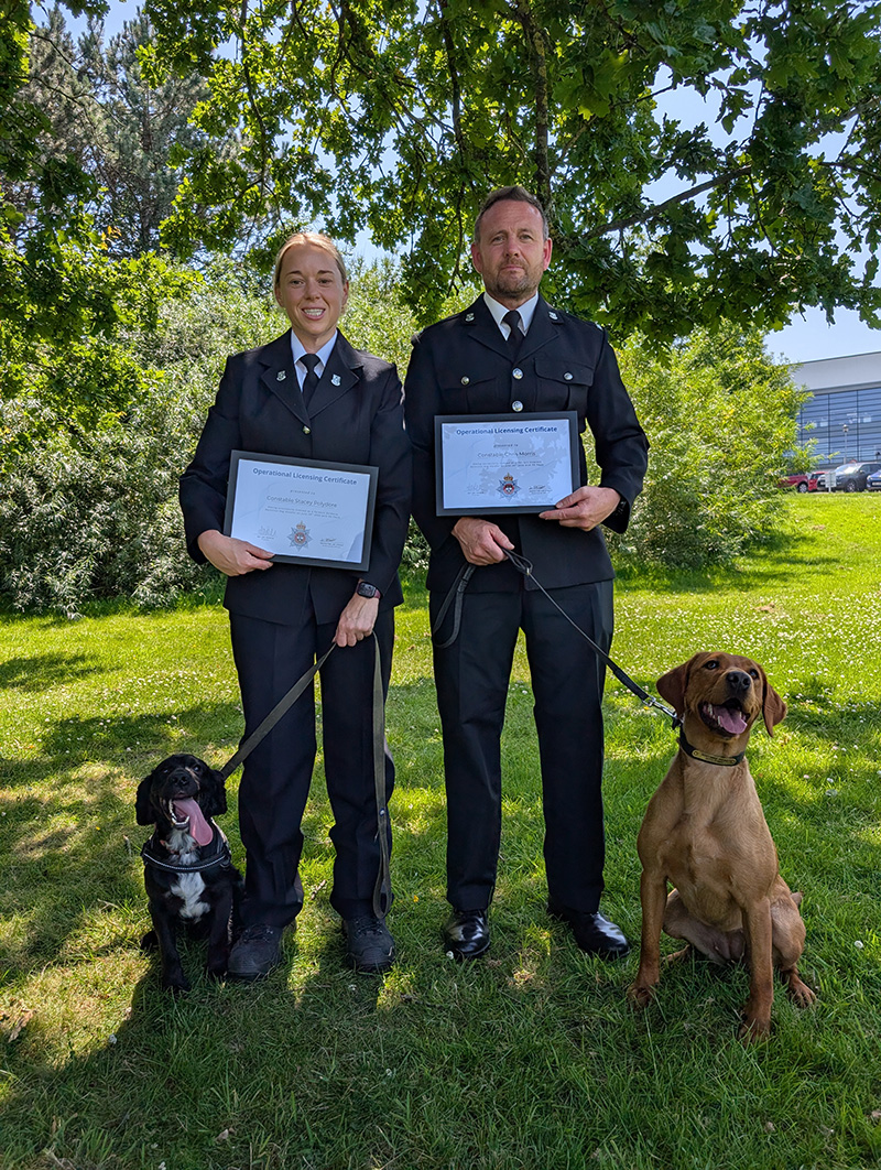 Two officers in uniform stand side by side with their dogs - a sandy coloured labrador and a black spaniel with a white chest