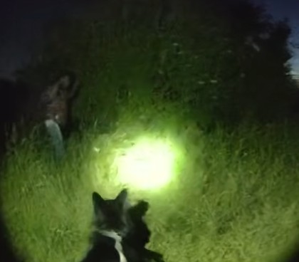 PD Yolo watches as a man walks with his hands up in a dark field lit by torchlight.