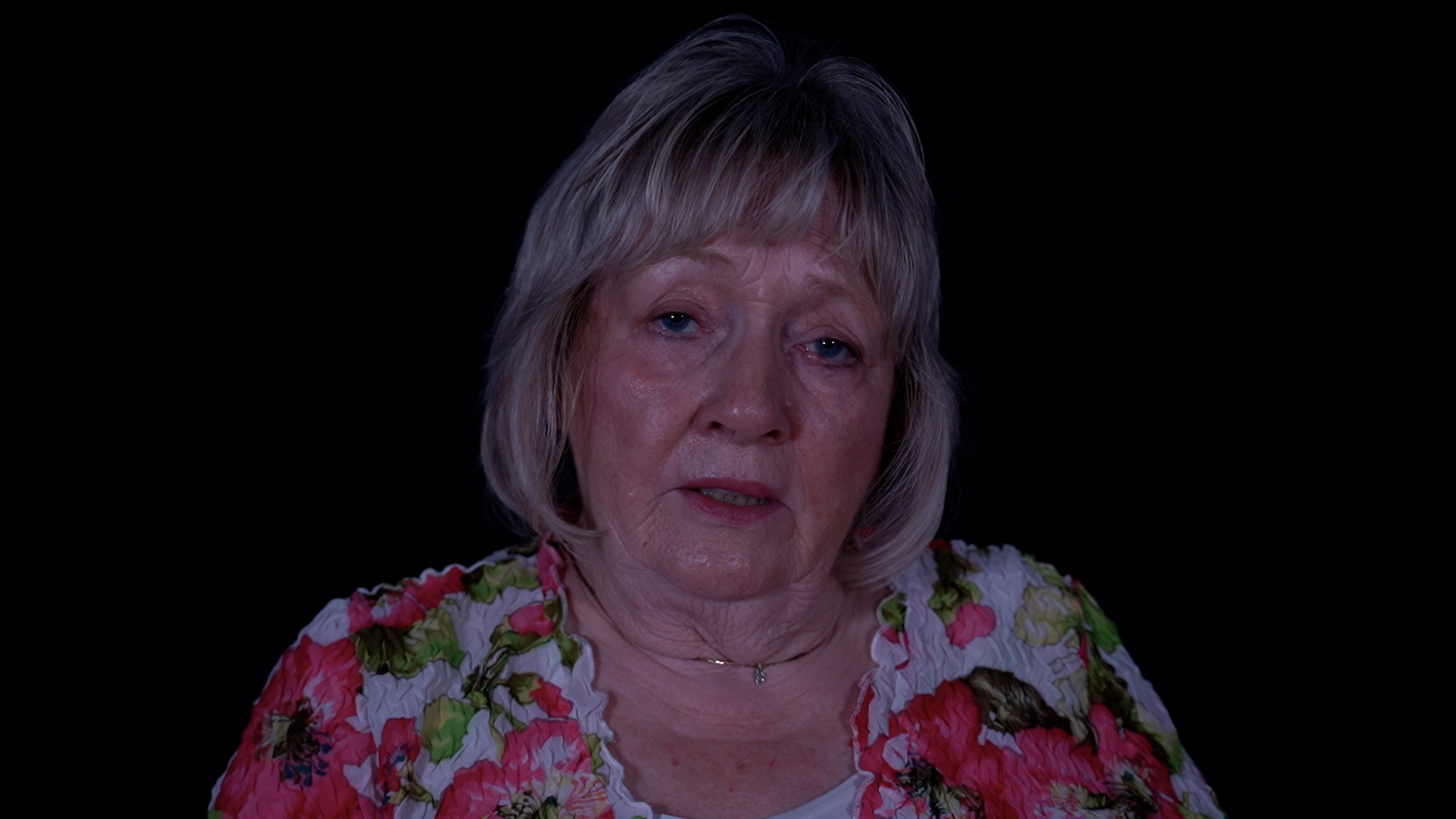 Angela Gaunt talking to the camera in a flowery top in front of a black background.
