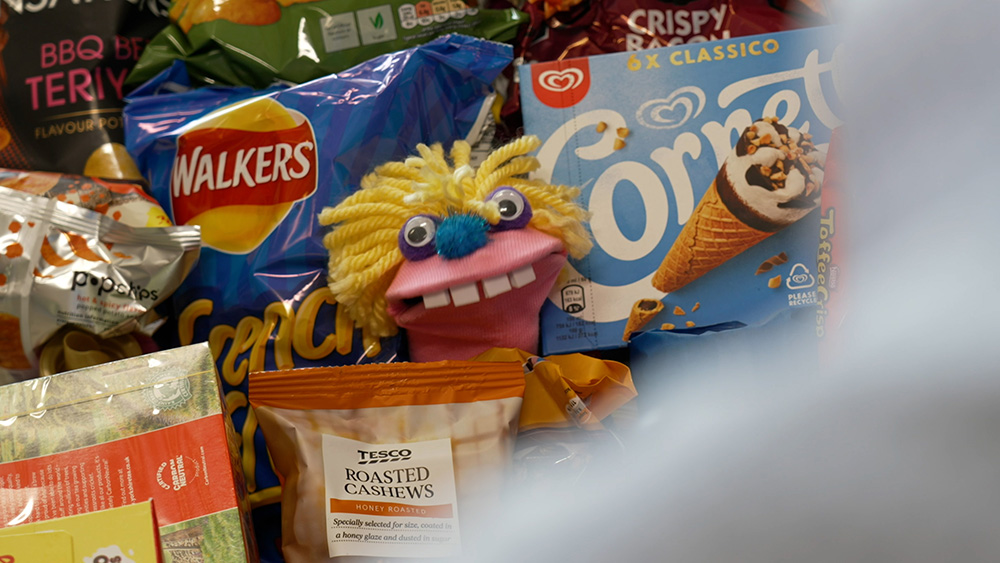 A bright pink sock puppet, with yellow hair, pops out out a full shopping trolley.